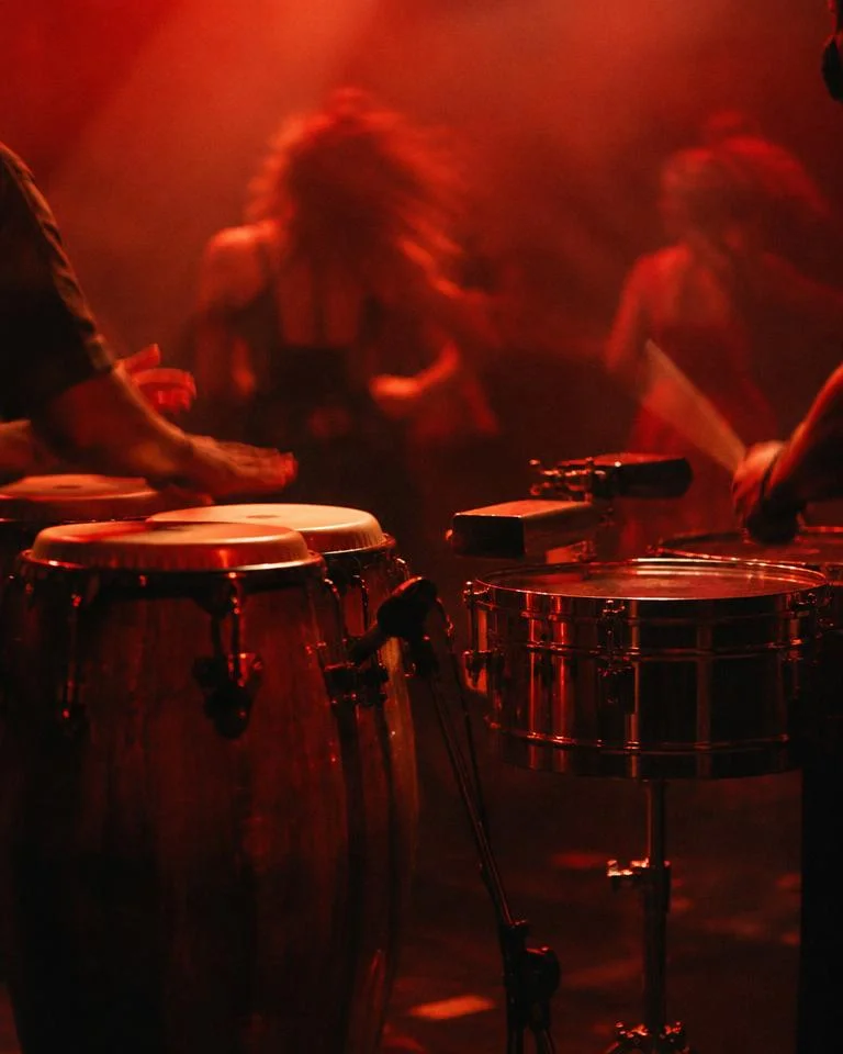 Congas and timbales in warm red light with dancers behind
